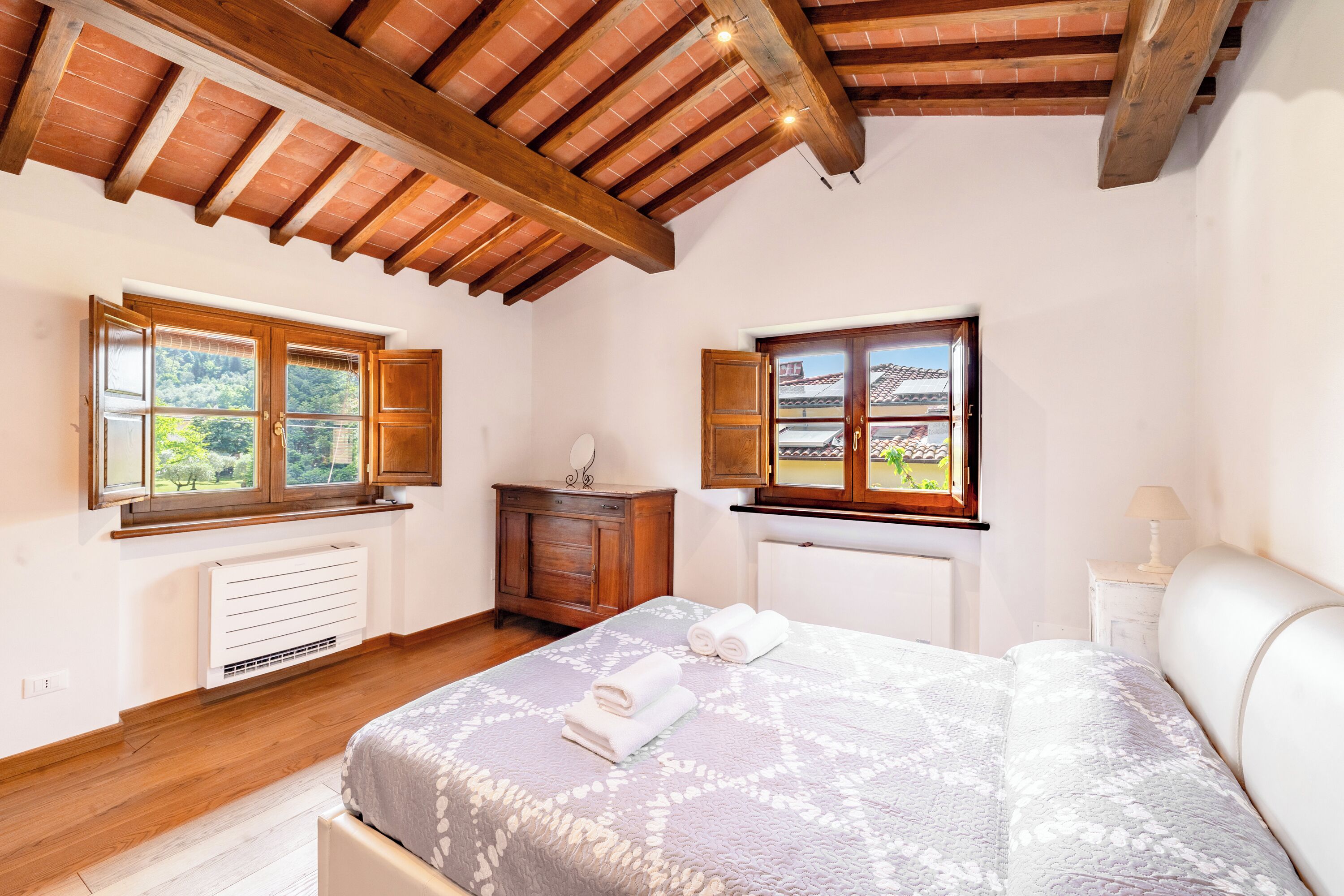 A bedroom with a bed, two windows, a radiator, wooden beams, and a white headboard.