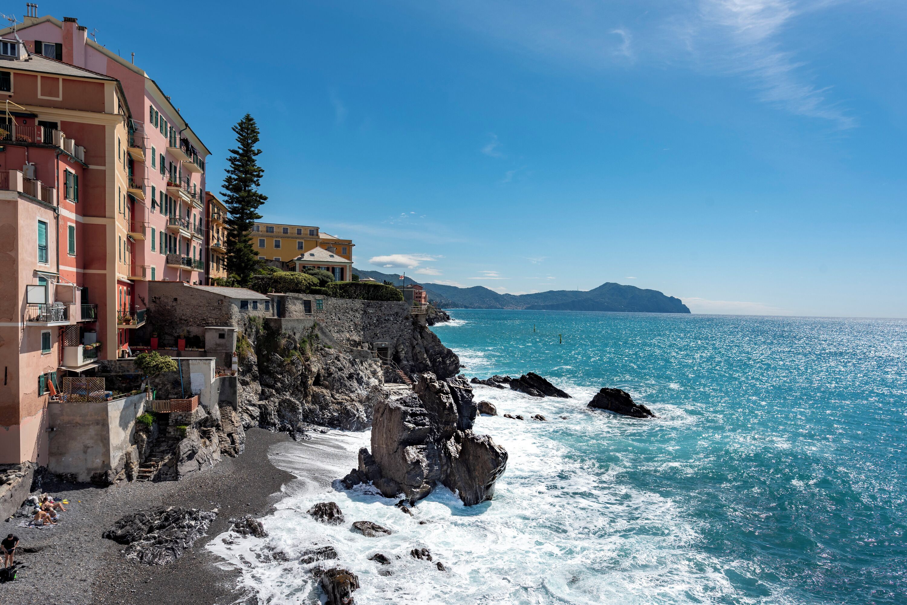 A coastal building with balconies overlooking the sea, a rocky shoreline, and a clear blue sky.