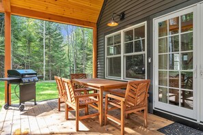 Eine Terrasse im Freien mit Holztisch und -stühlen, einem Grill und einem großen Fenster mit Blick auf Bäume.