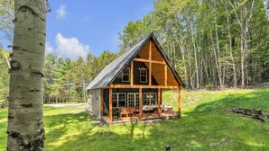 Una cabaña con un porche, terraza de madera y un gran ventanal con vista a un bosque.