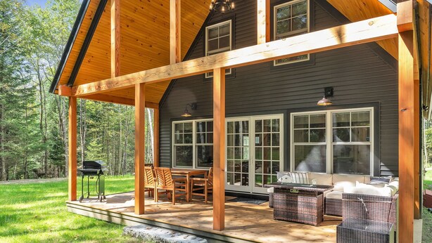 Una terraza de madera con mobiliario exterior, una parrilla y un área de estar cubierta con ventanas grandes.