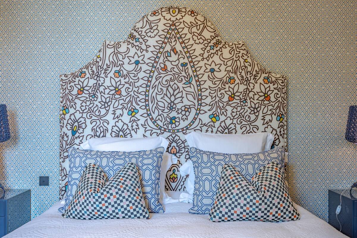 Ein Bett mit gemusterten Bettbezügen und Kissen, ein Kopfteil mit floralem Muster und eine Wand im Hintergrund mit Musterung.