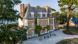 A stone building with multiple windows and a sloped roof, surrounded by trees and a gravel driveway. - Villa Haute Guais  (Dinard)