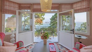 A room with a large window, a hanging light fixture, a table with a vase of flowers, and patterned curtains. - Villa Haute Guais  (Dinard)
