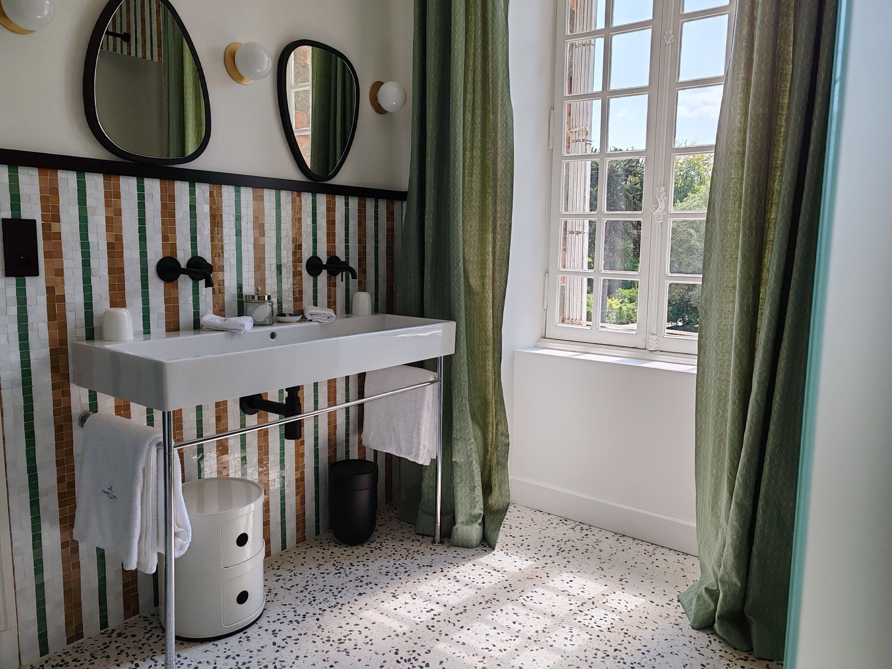 A bathroom with a white sink, two mirrors, a towel rack, and a window with green curtains.