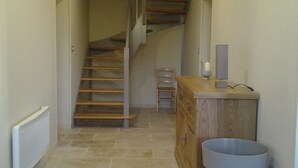 A narrow hallway with a wooden staircase, a radiator, a wooden cabinet, and a trash bin. - Gîte des 3 Puits (Caumont)