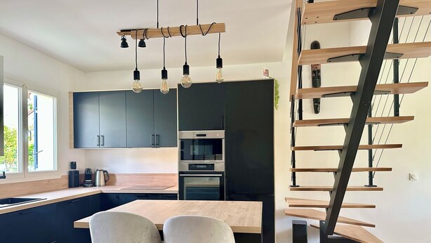 Une cuisine moderne avec des meubles de couleur sombre, un îlot central en bois et un four encastré. On y trouve deux tabourets de bar et une machine à café sur le comptoir.