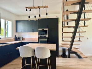 A modern kitchen with dark cabinets, a wooden island, and a built-in oven. There are two bar stools and a coffee machine on the counter.