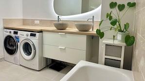 Une salle de bain moderne équipée d’un lave-linge intégré, d’un grand miroir ovale, d’un lavabo sur colonne et d’une plante en pot.