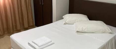 Una cama bien hecha con sábanas blancas y una toalla doblada, un armario marrón y un aire acondicionado de pared.