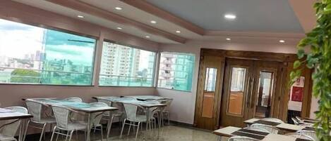 A dining area with tables and chairs, large windows with a city view, and a ceiling with recessed lighting.