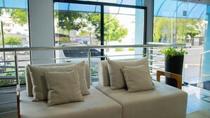 Un hall d’hôtel moderne avec un espace salon comprenant deux canapés aux coussins beiges, une plante en pot et une vue sur la rue grâce à de grandes fenêtres.