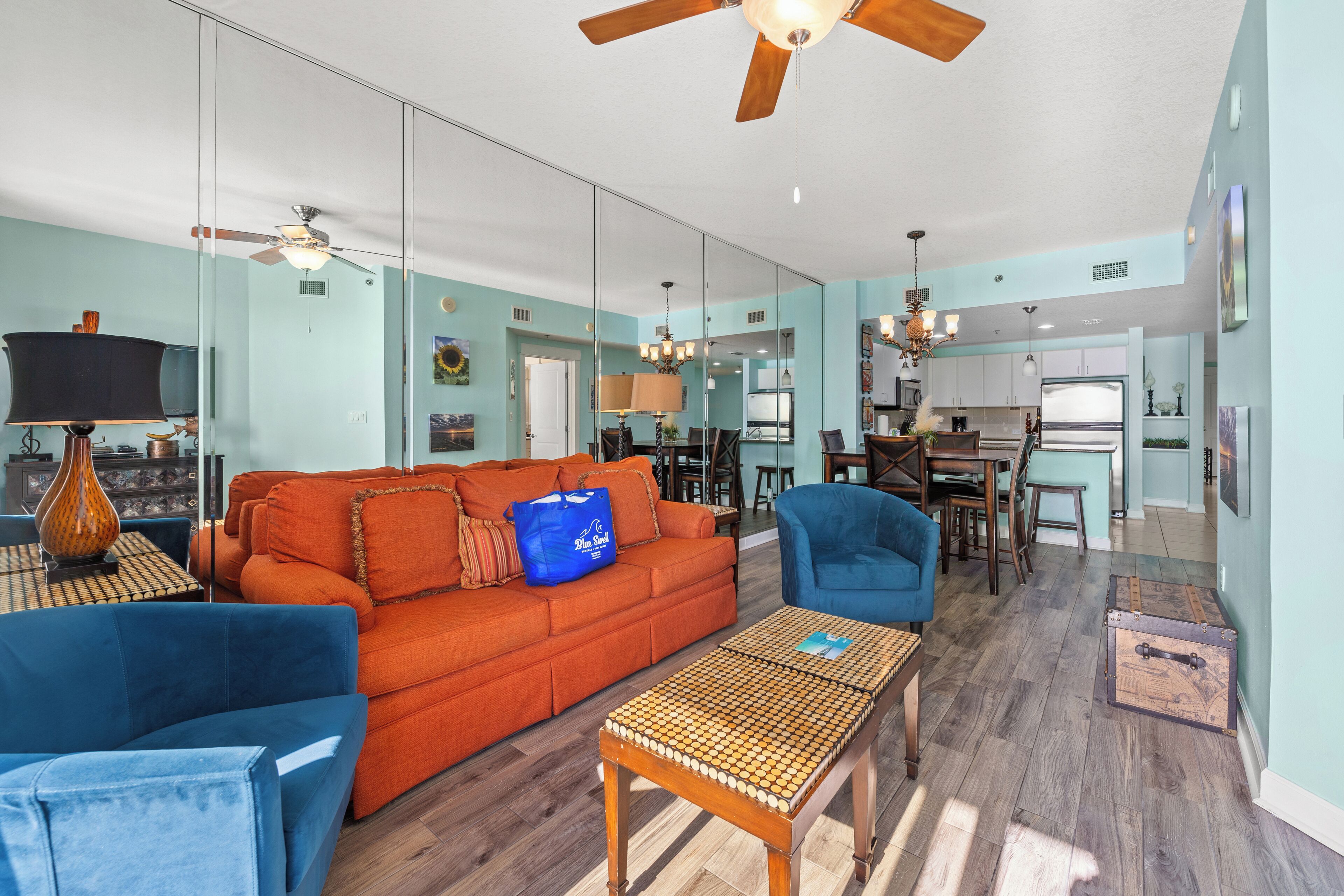 A spacious living room with a large orange sofa, blue armchairs, a wooden coffee table, and a ceiling fan. A kitchen is visible in the background.