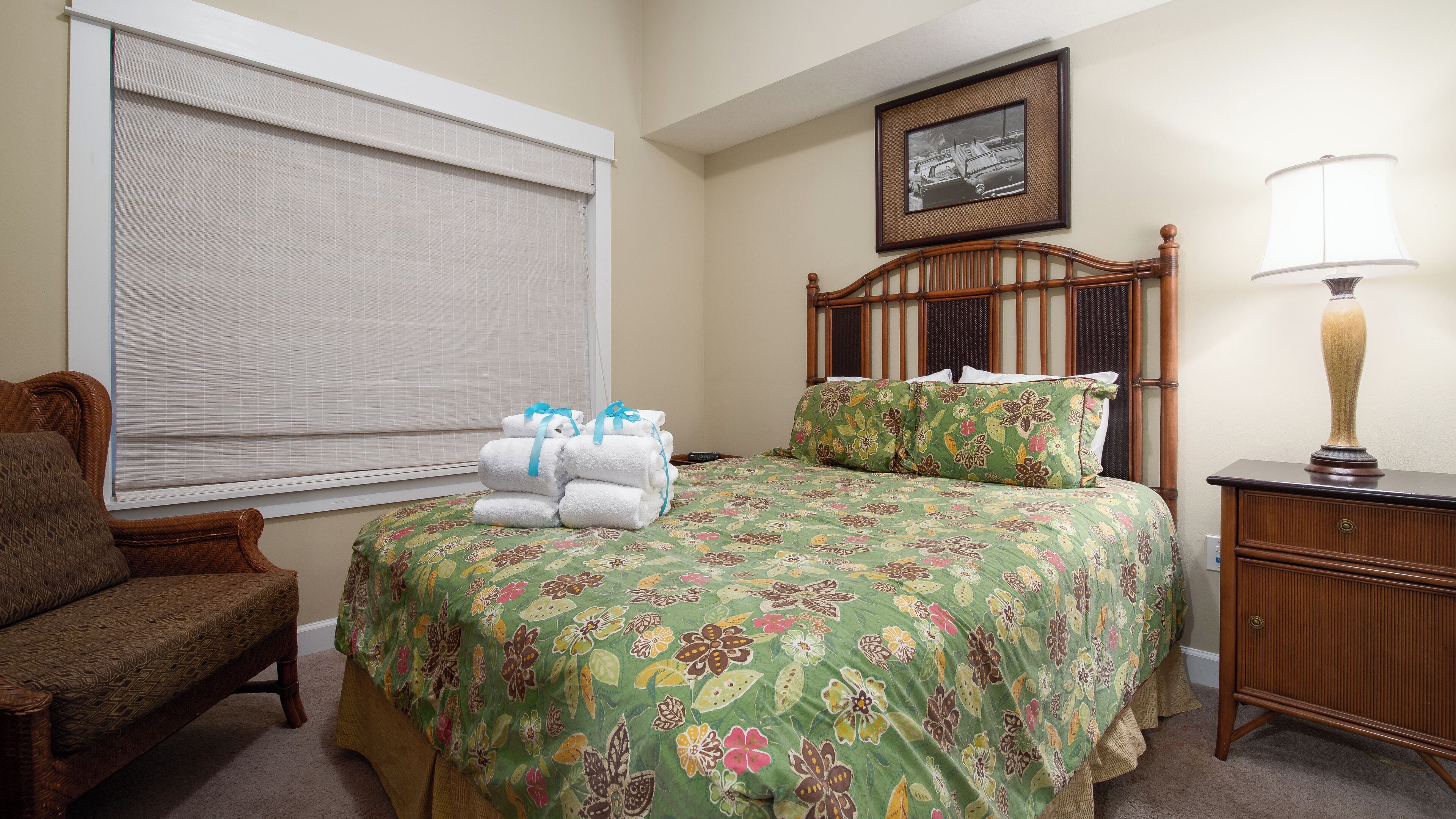 A hotel room with a bed, a chair, a nightstand, a lamp, a window with blinds, and a framed picture on the wall.