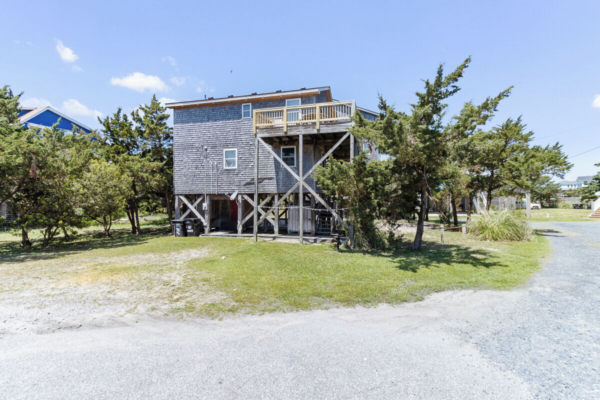 Un bâtiment en bois à deux étages, doté d’une terrasse au niveau supérieur, entouré d’arbres et bordé d’une allée en gravier.
