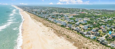 An aerial view of a coastal residential area with a beachfront, houses, and a pier extending into the ocean.