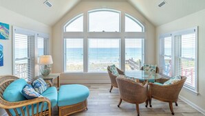 A sunlit room with a large arched window, wicker furniture, and a glass table. - Beautiful oceanfront home in Duck with gorgeous views! (Duck)
