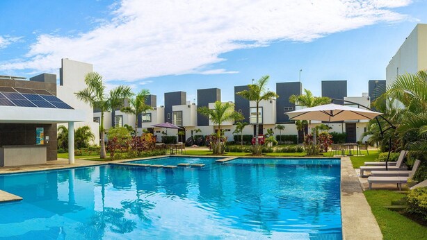 Una piscina con sillas de descanso y un área de estar cubierta, rodeada de palmeras y edificios modernos.