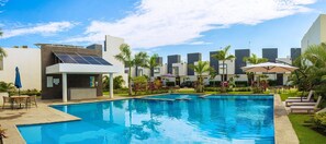 Una piscina con sillas de descanso y un área de estar cubierta, rodeada de palmeras y edificios modernos.