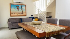 Un comedor con una mesa de madera, sillas grises y un mantel decorado. Un sofá gris con cojines está apoyado contra la pared, y hay un cuadro de un elefante encima.