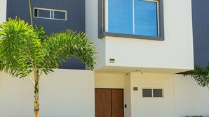 Un moderno edificio de dos pisos con un gran ventanal en el segundo piso, una puerta de madera y un pequeño jardín con un árbol y arbustos.