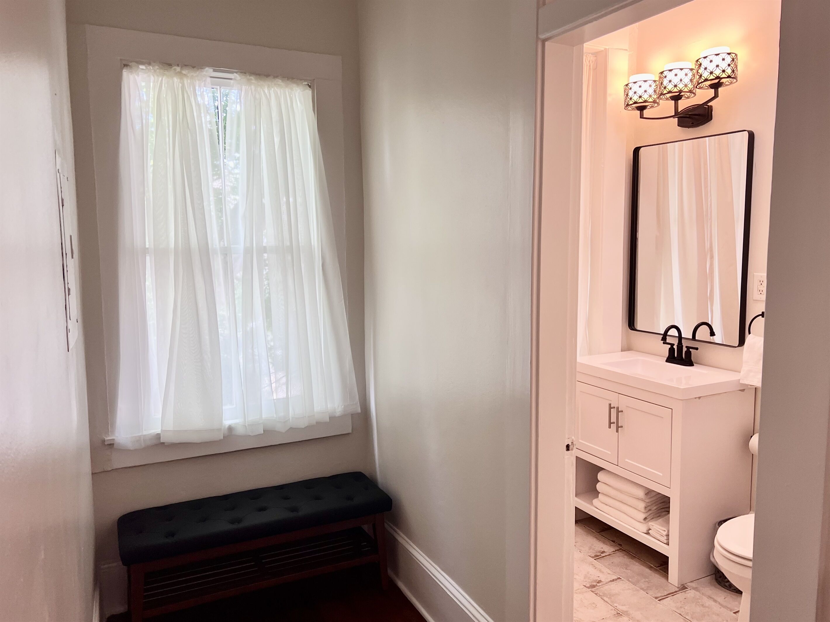 Une salle de bain avec une fenêtre, un banc, un meuble-lavabo avec miroir et des toilettes.