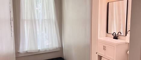 A bathroom with a window, bench, vanity with mirror, and toilet.