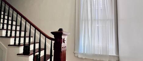 Una escala de fusta amb barana i pomes de canya, que condueix a un espai amb un gran finestral cobert per cortines lleugeres.