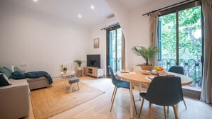 Una sala de estar moderna con sofá, mesa de comedor con sillas, televisor y un ventanal con cortinas.