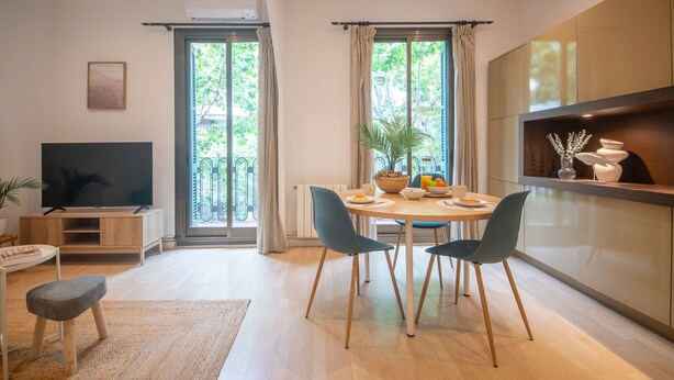 Una sala de estar moderna con televisor, mesa de comedor, sillas y sofá. Hay ventanas amplias con cortinas, una planta y una estantería con adornos.