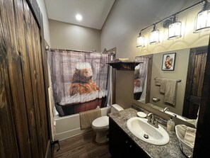 Een badkamer met een houten vloer, een douche met gordijn met beer thema, een wit toilet, een wastafel met granieten blad en een spiegel met verlichting erboven.