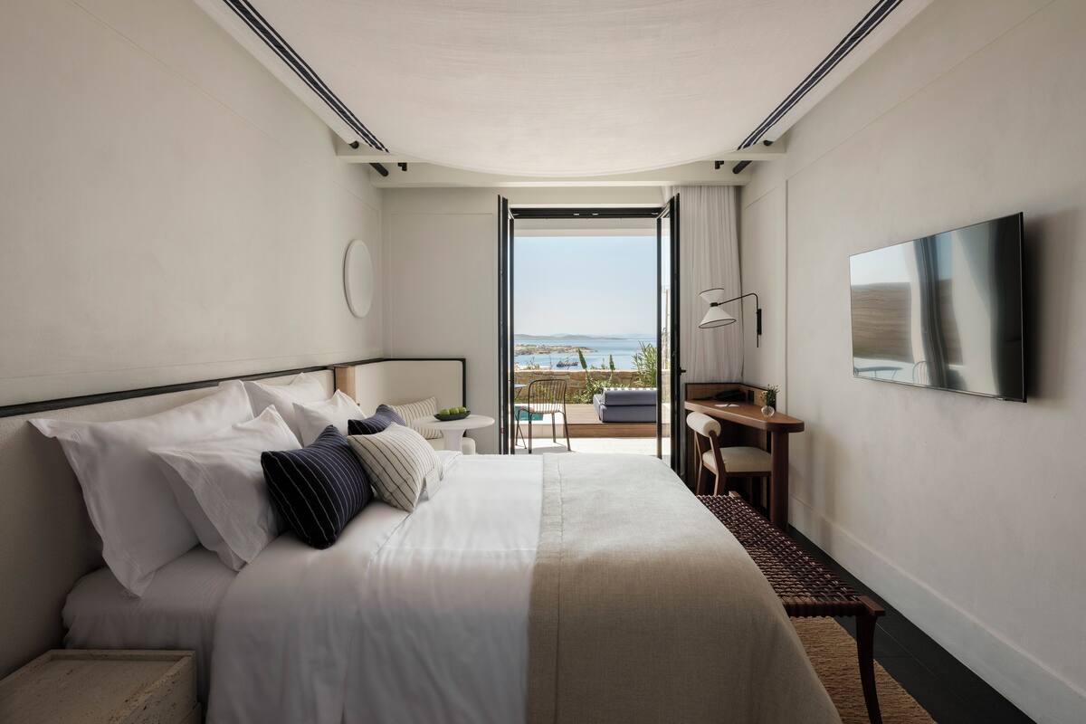 A hotel room with a large bed, a desk, a chair, a TV, and a balcony with a view of the sea.
