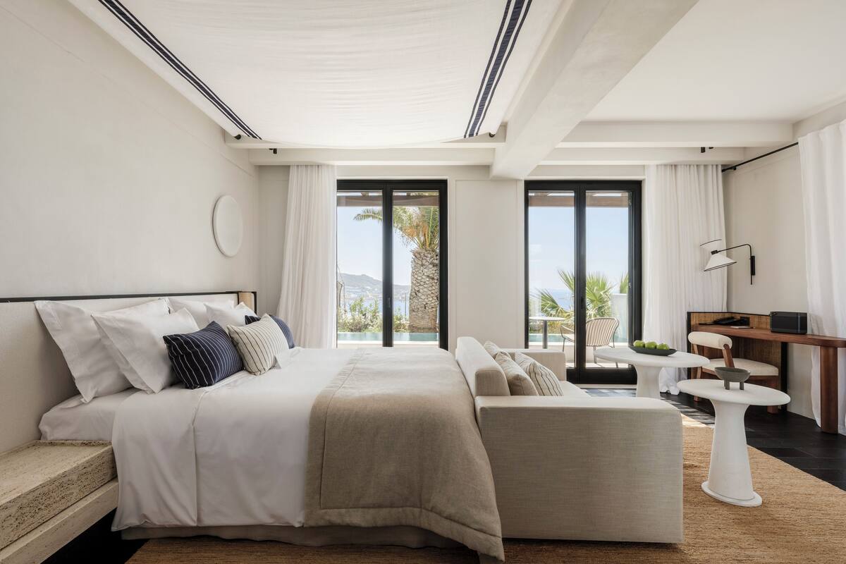 A modern bedroom with a large bed, a sofa, a small table, and a view of the outdoors.