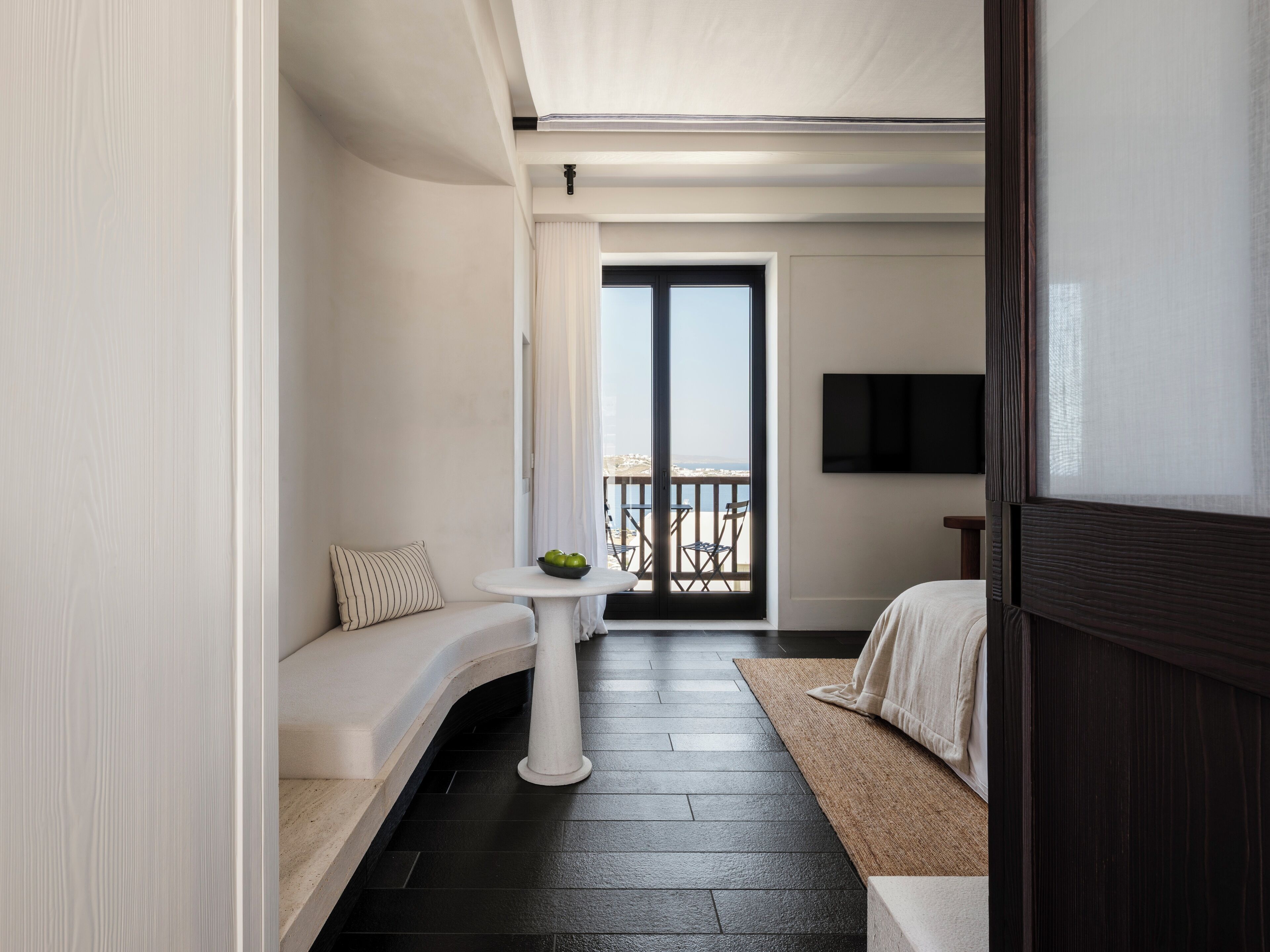 A modern hotel room with a curved sofa, a small round table with a bowl of fruit, a bed with a dark headboard, and a balcony with a view of the sea.