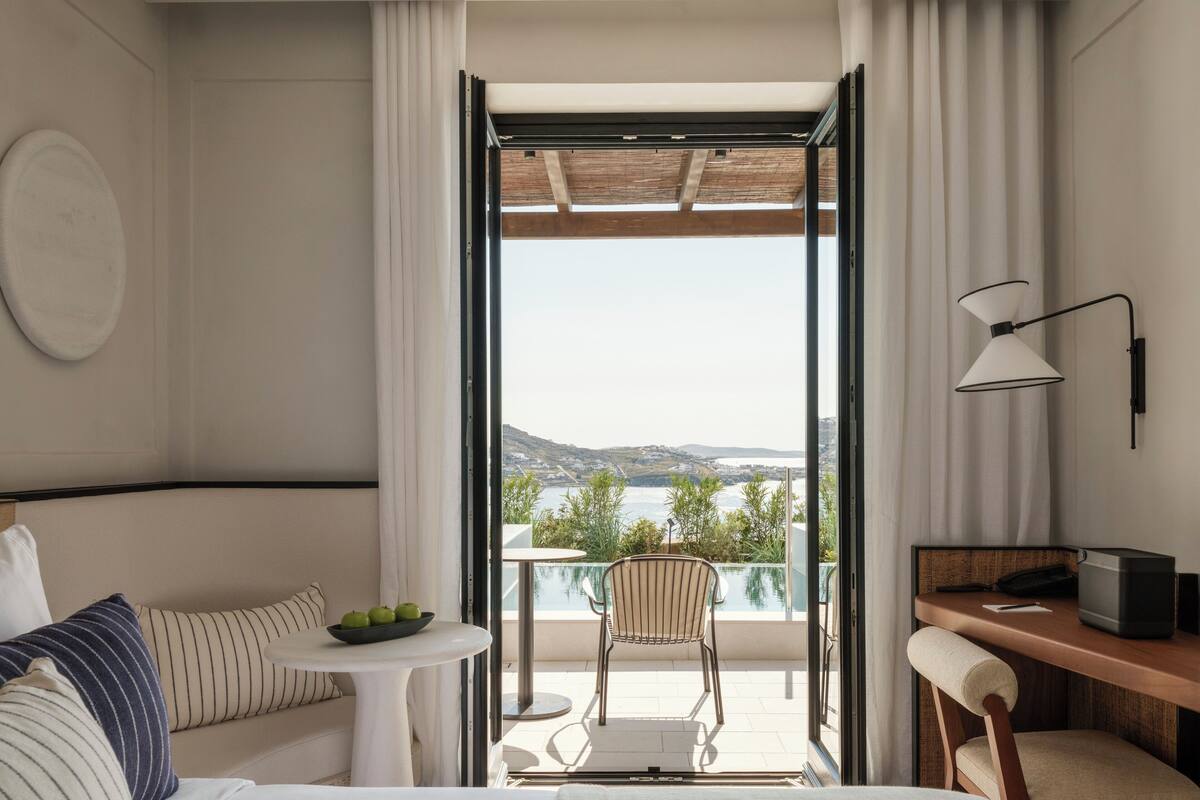 A hotel room with a bed, a small table with apples, a chair, a desk with a chair, and a view of the sea and hills.