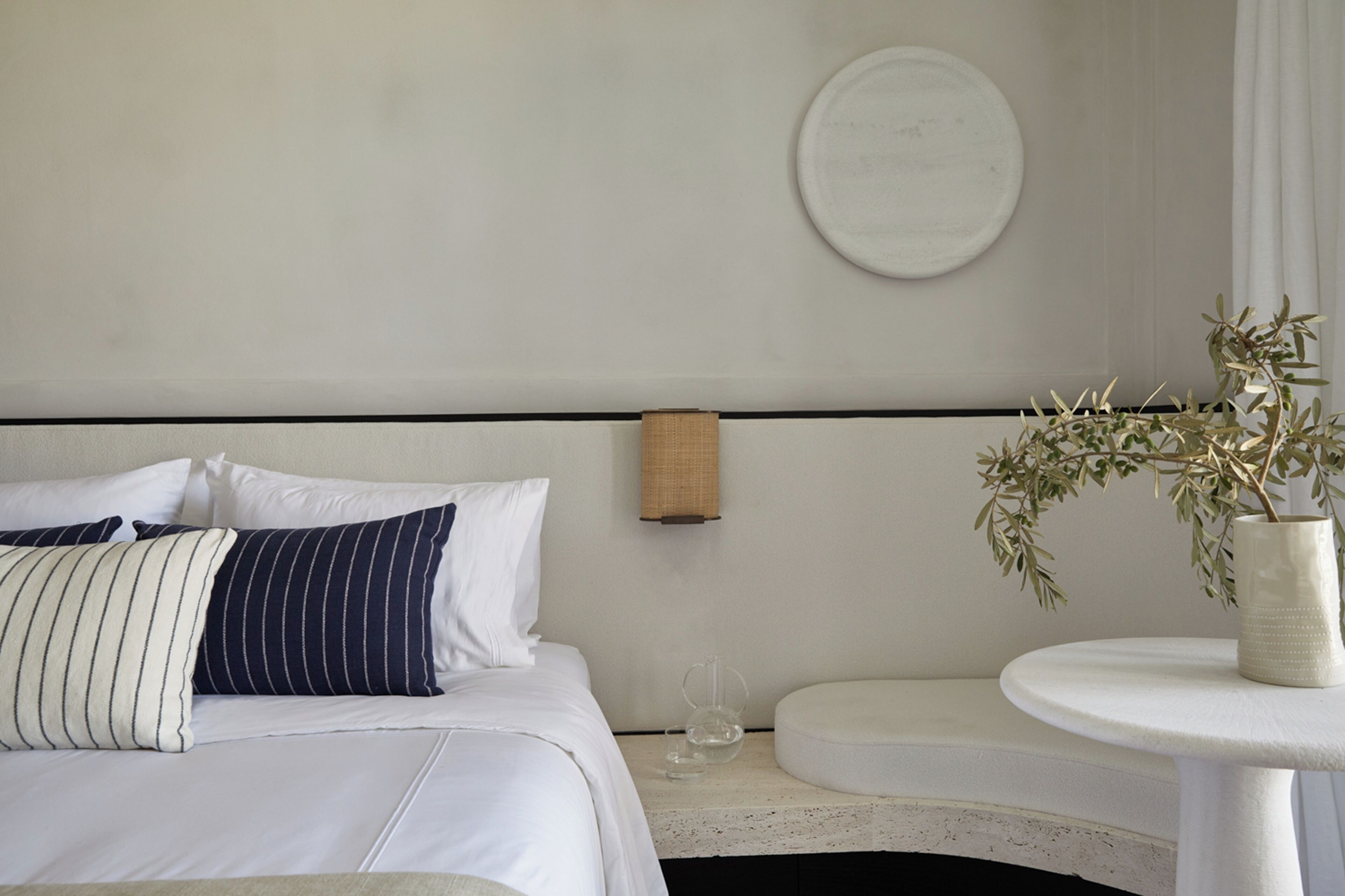 A hotel room with a bed, a bedside table with a vase of plants, a wall-mounted lamp, and a round mirror.