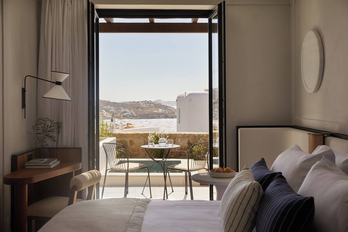 A hotel room with a bed, a desk, a chair, a small table with a bowl of fruit, and a view of the beach and mountains through an open door.