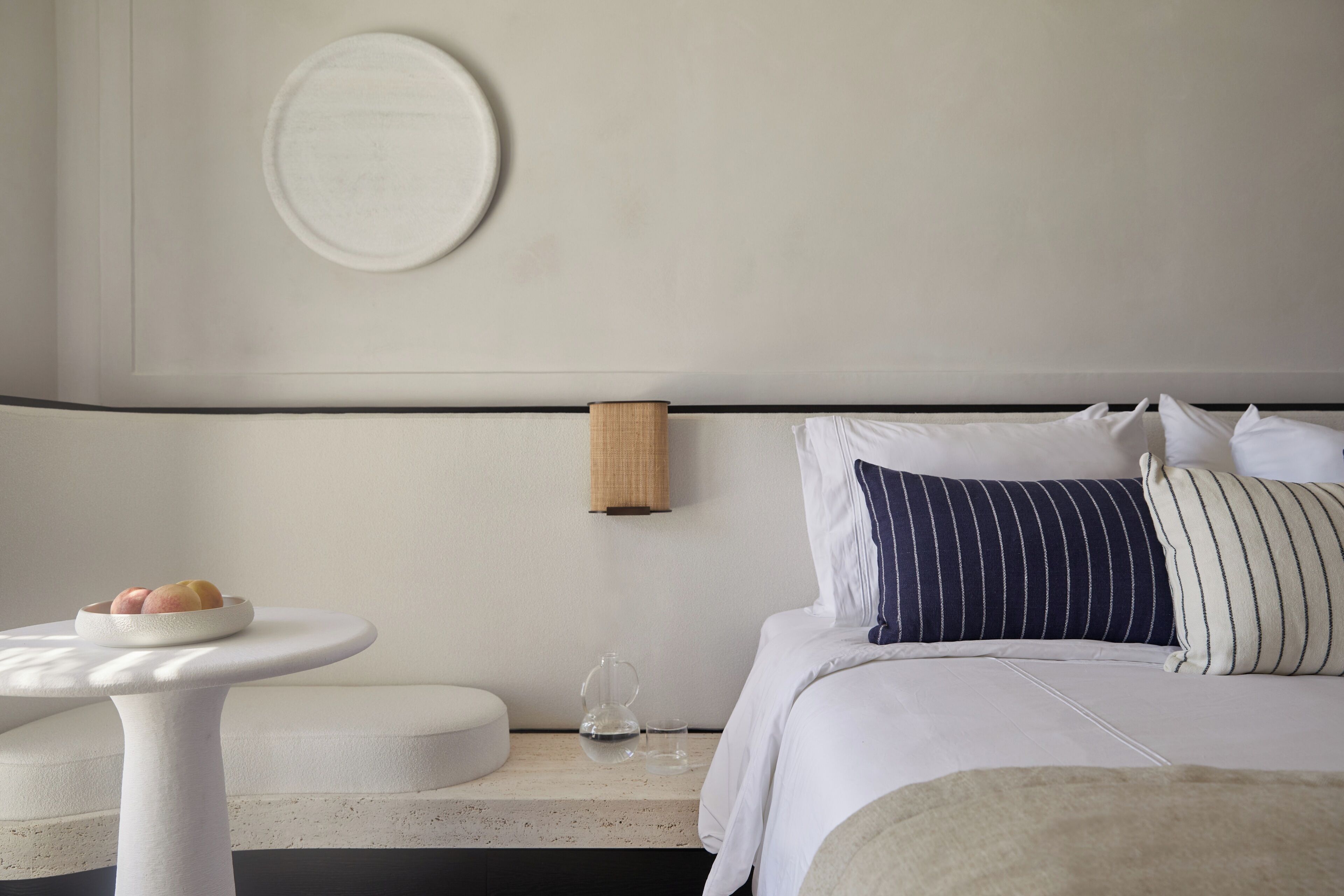 A modern hotel room with a bed, a round table with a plate of fruit, a wall-mounted lamp, and a round mirror.