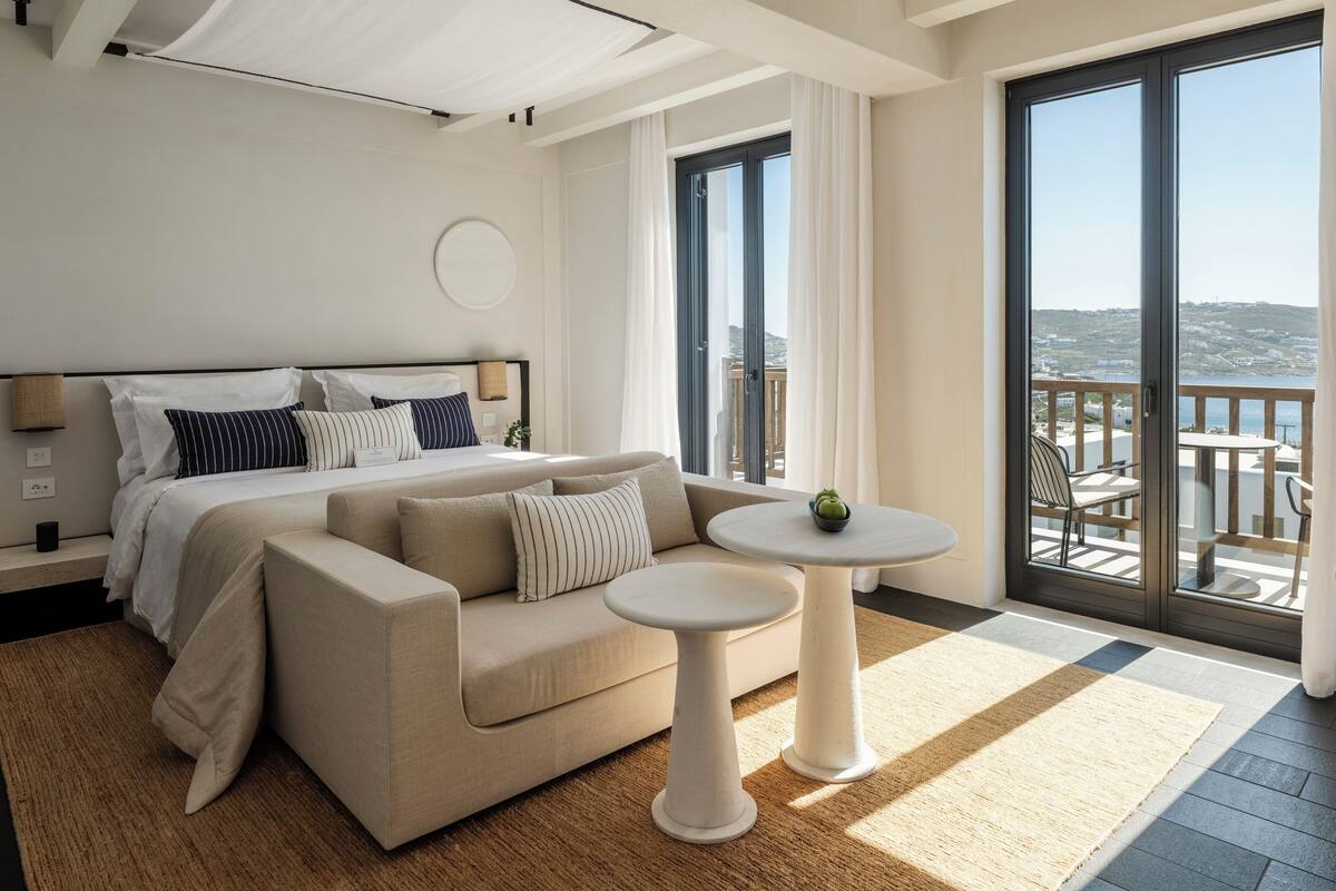 A modern hotel room with a large bed, a sofa, a small table, and a balcony with a view.
