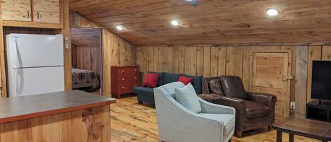 Een gezellig interieur van een chalet met houten plafond, een plafondventilator, een koelkast, een bank, een leren fauteuil, een kleine tafel en een televisie.