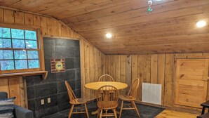 Et hyggeligt værelse med trævægge og flisegulv, med et rundt træbord med stole, en sofa og et vindue med udsigt til træer.
