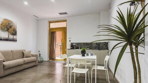 A modern living room with a beige sofa, a dining table with white chairs, a kitchen area, and a large potted plant.