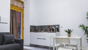Ein modernes Wohnzimmer mit grauem Sofa, einem Esstisch mit weißen Stühlen, einer Küchenzeile mit Musterfliesen und einem Topfpflanzen.