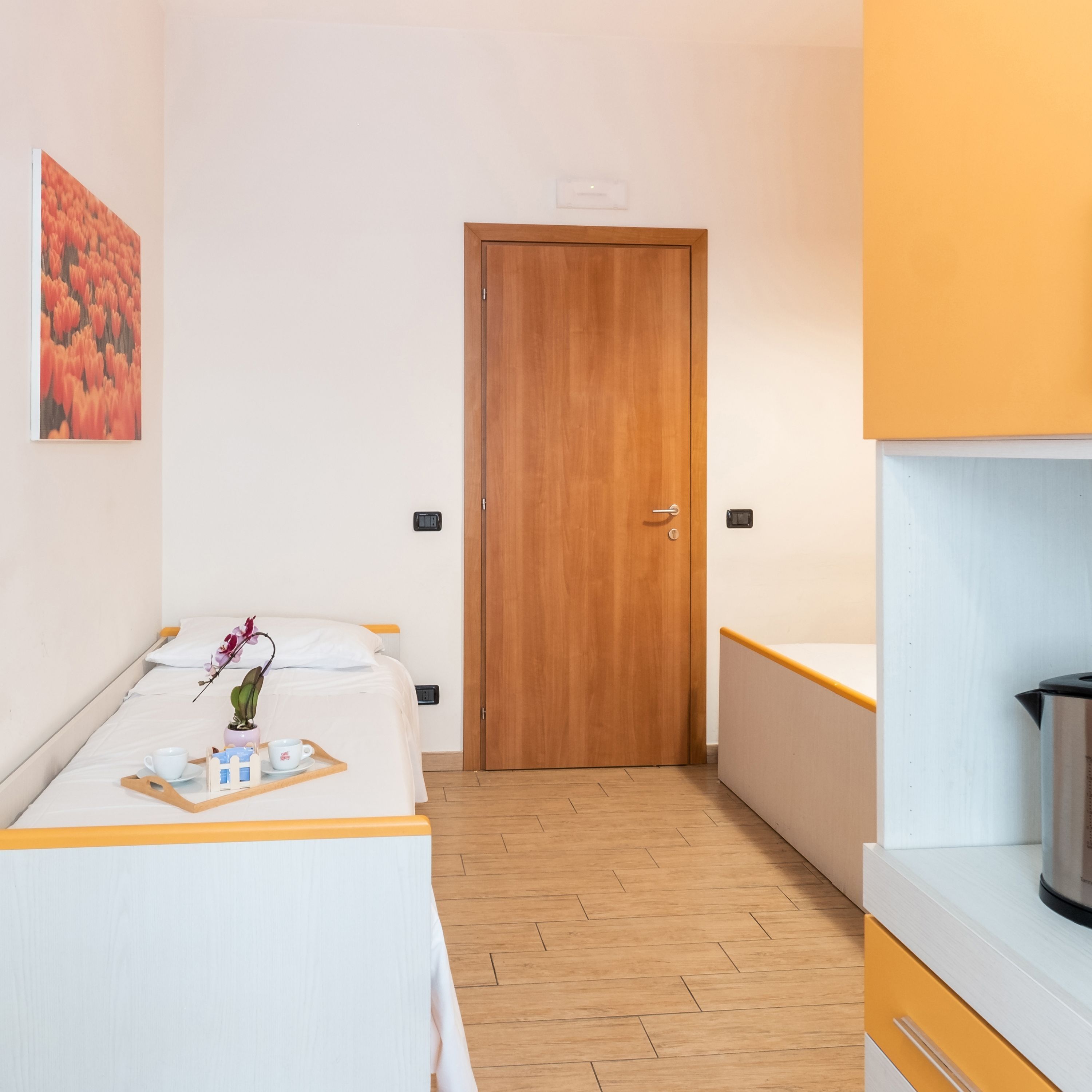 Une chambre d’hôtel avec une porte en bois, un lit sur lequel se trouve un plateau avec des articles pour le petit-déjeuner, une petite plante en pot et un tableau accroché au mur.