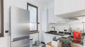 Una cocina moderna con un refrigerador de acero inoxidable, un fregadero y una estufa. Hay una ventana con persianas, un bote de basura y una pequeña planta en maceta sobre la encimera.