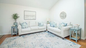 Ein Wohnzimmer mit zwei weißen Sofas, einem großen Teppich mit Muster, einem Beistelltisch mit Lampe und einer kleinen Pflanze.