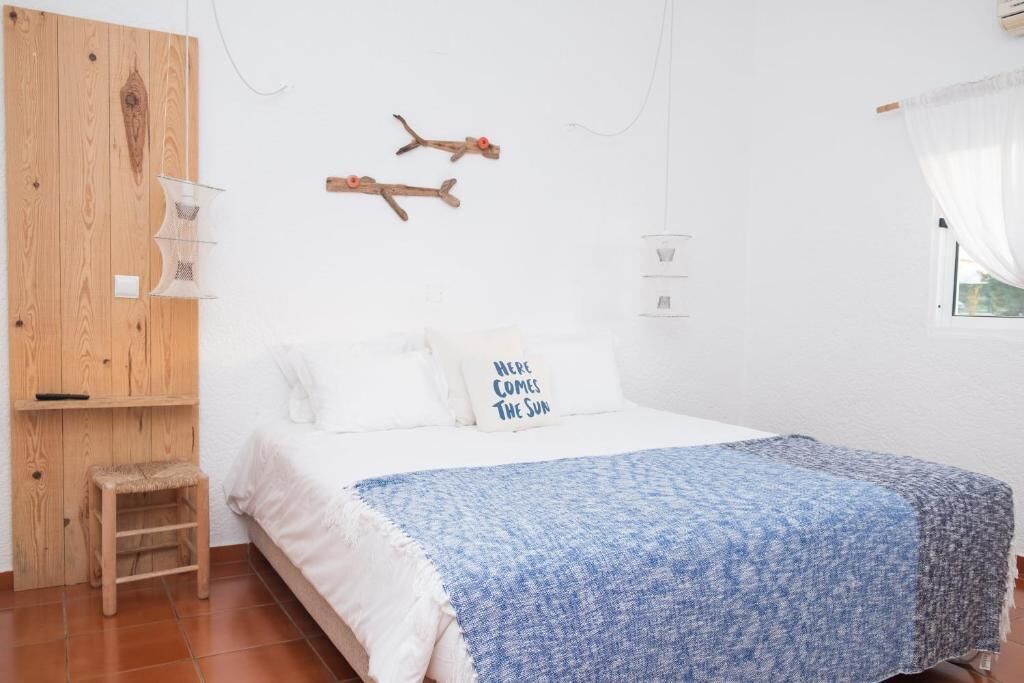 A bedroom with a bed, a wooden wardrobe, a small stool, and a wall-mounted shelf.