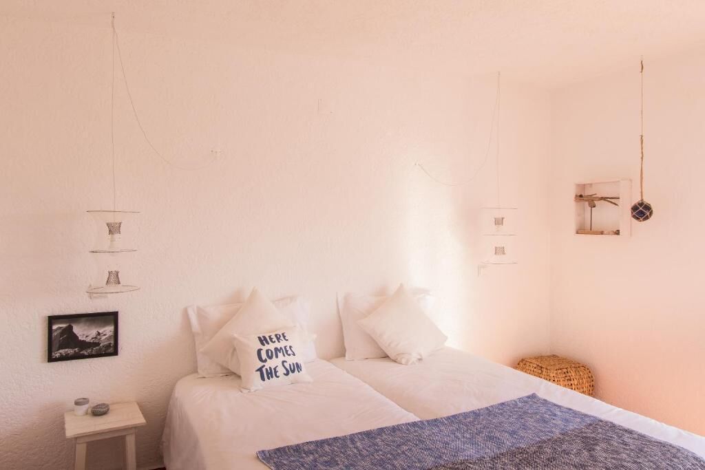 Une chambre à coucher comprenant un lit, des oreillers, une table de chevet, une étagère fixée au mur, une lampe suspendue et un petit tabouret en osier.