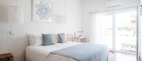 Quarto com uma cama grande, uma mesinha e uma cadeira. Há uma janela com vista para o exterior.