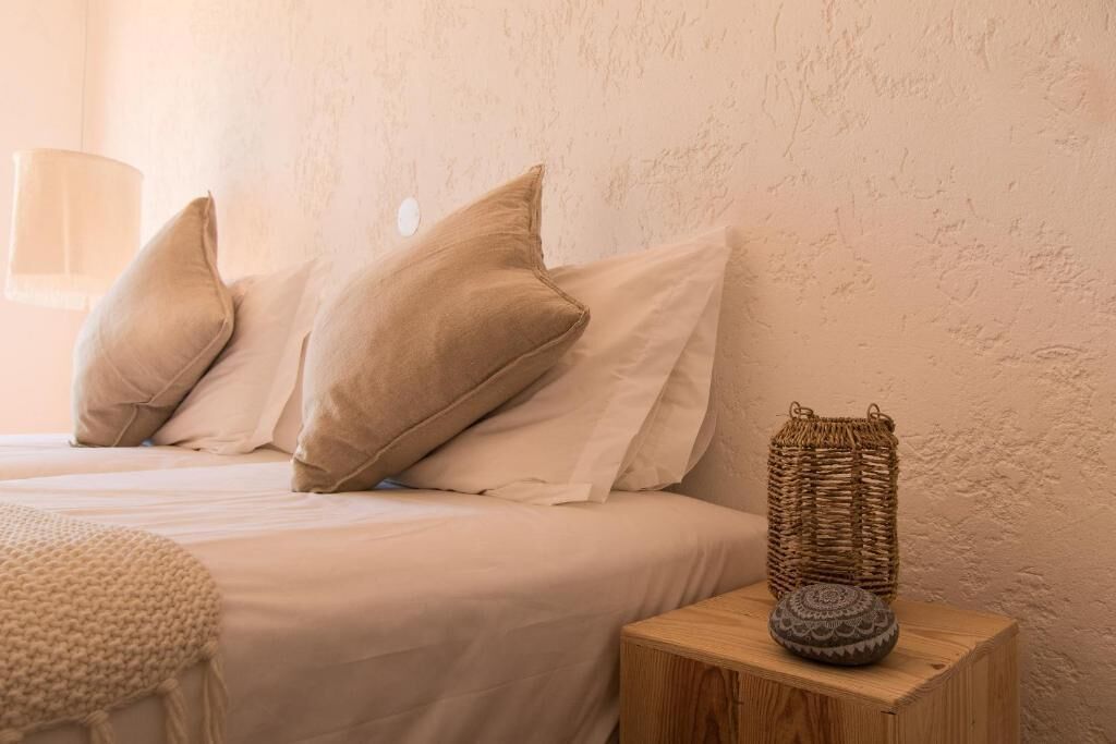 Una cama con ropa de cama y almohadas color beige, una cesta tejida con un objeto estampado sobre una mesita de noche de madera, y una pared con textura.